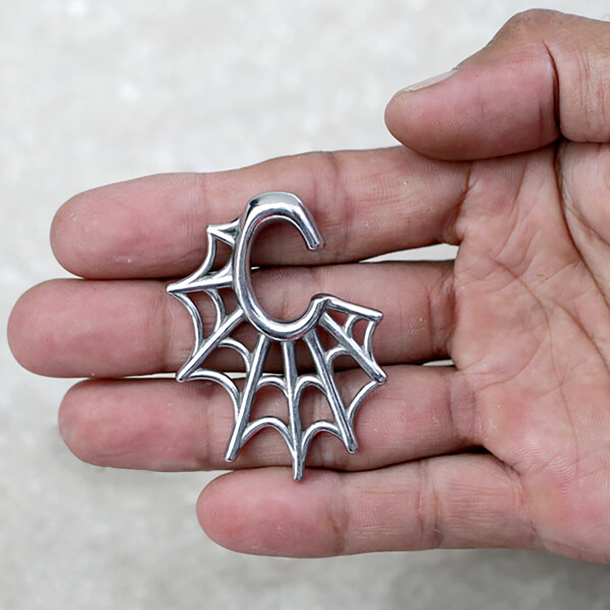 A hand holding a silver keychain shaped like a spider web, reminiscent of the intricate design of the Spider Web Gothic Steel Ear Hangers in Silver | 2 gauge by Kaya Movement. The keychain features a hook at the top for attaching to keys or a bag. The background is a light, neutral color.